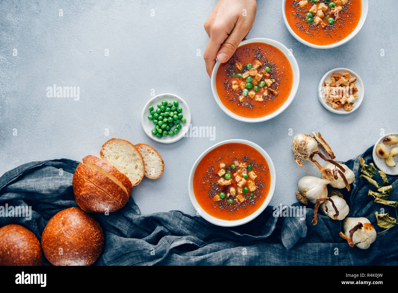 Vegan tomato soup served in three white bowls accompanied by bread rolls, roasted garlic, croutons and peas. A hand is grabbing one bowl. Stock Photo