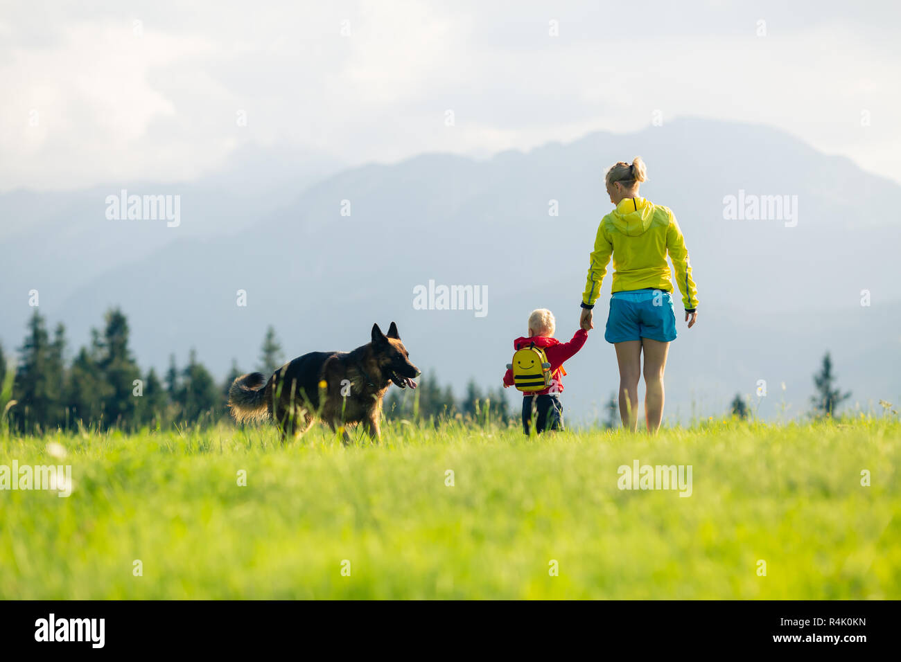 Mother with baby boy walking a dog on green meadow. Hiking adventure with child on summer family trip in mountains. Vacations or weekend activity with Stock Photo