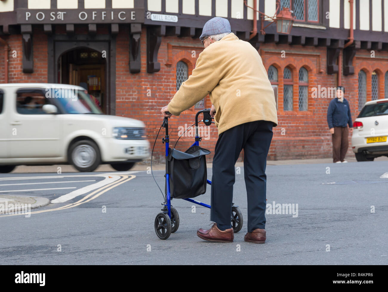 Elderly man walking across a road assisted by a 3 wheeled walker in the UK. Senior person shopping with wheeled walking frame. Stock Photo