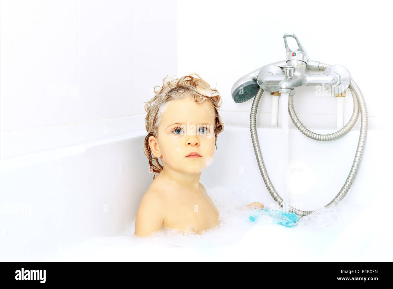 Cute adorable baby taking bath in washing sink on water tap background. Little healthy girl with big blue eyes having fun and playing with soap foam.  Stock Photo