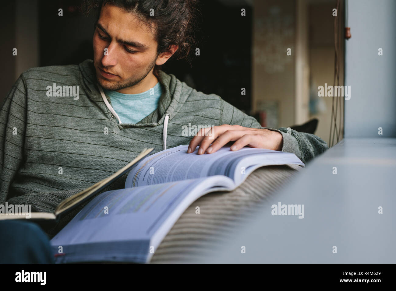 Student making notes from a reference book sitting on a couch. Young man studying seriously sitting at home. Stock Photo