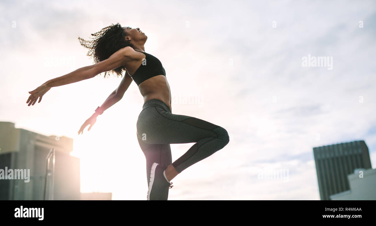 Woman in fitness wear jumping high standing on rooftop with sun flare in the background. Female athlete doing exercises in the morning on terrace of a Stock Photo