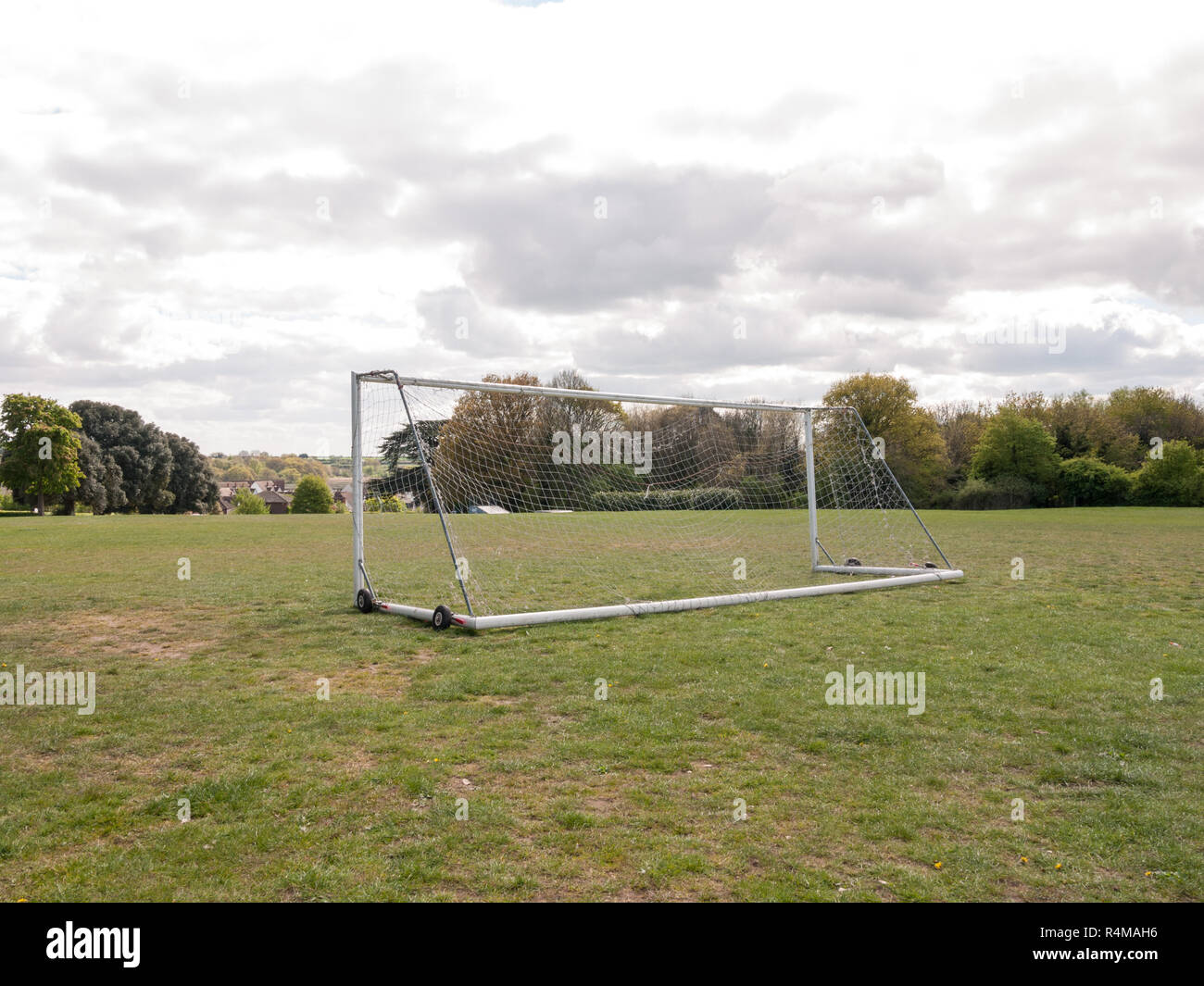 An Empty and Unused Goal Post with A White Net in the Middle of A Park with Grass and Soil on the Ground, Wheels on the Frame to Help Move it and Houses and Trees in the Background, Used For Football and for A Goal Keeper No Person Seen From Behind Stock Photo