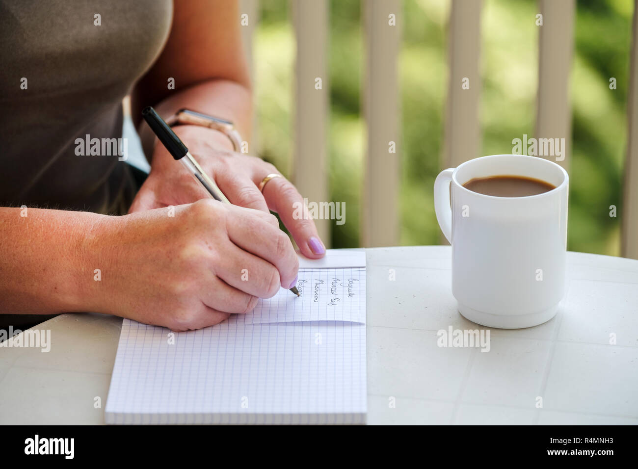 A woman sat at a table, with a cup of coffee, writing a shopping list before visiting the shop to get food and provisions Stock Photo