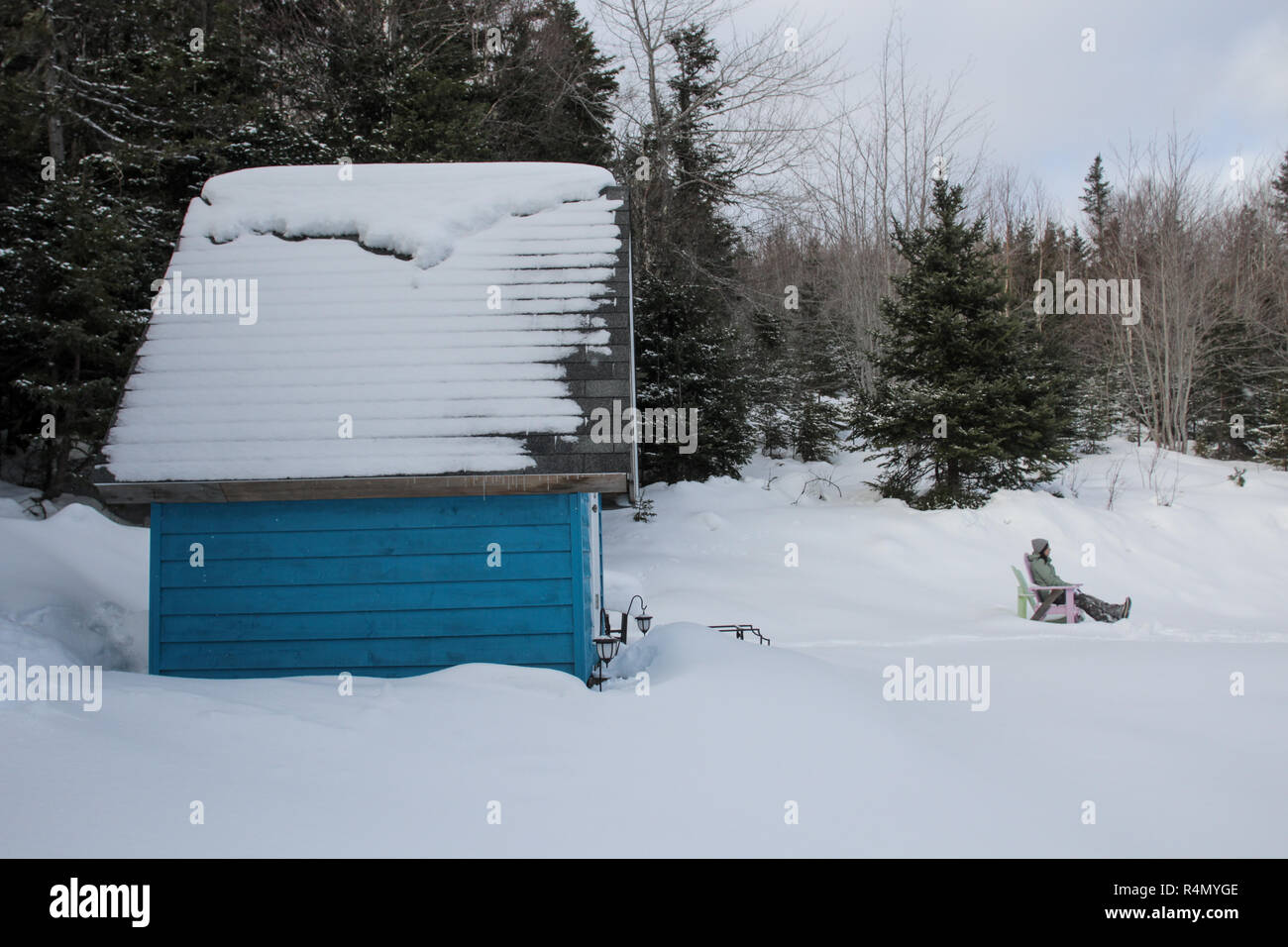 A woman relaxing in an adirondack chair in front of her blue tiny home in the snow Stock Photo