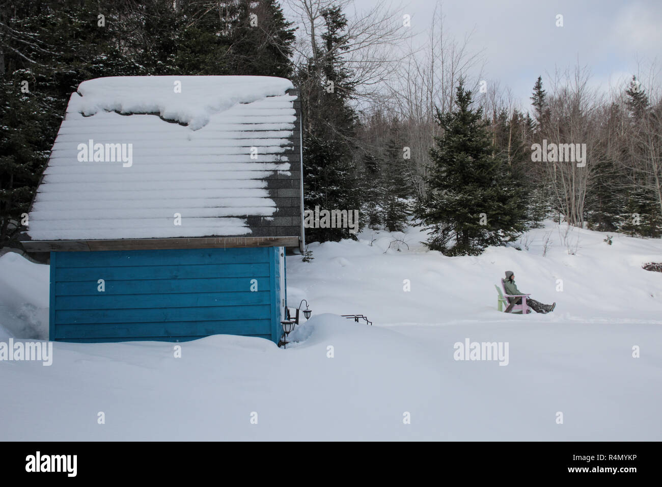 A woman relaxing in an adirondack chair in front of her blue tiny home in the snow Stock Photo