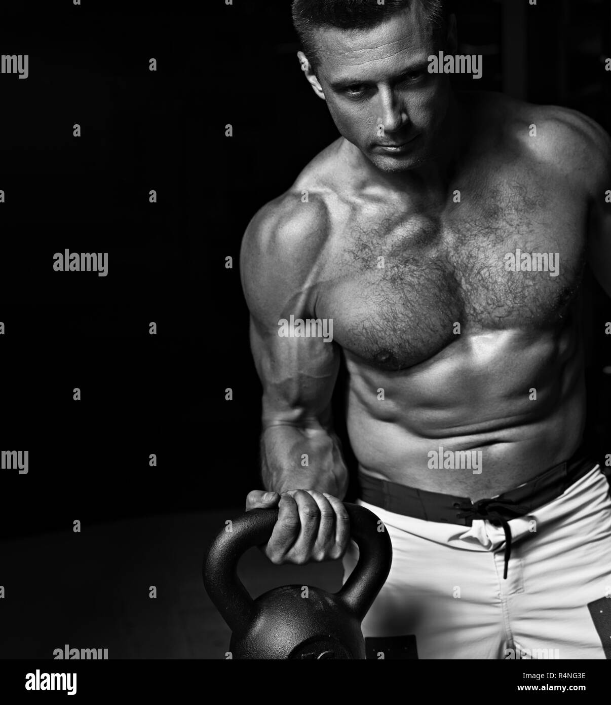 Strong angry athletic man doing the exercises with kettlebells on dark shadow background. Closeup portrait. Black and white Stock Photo