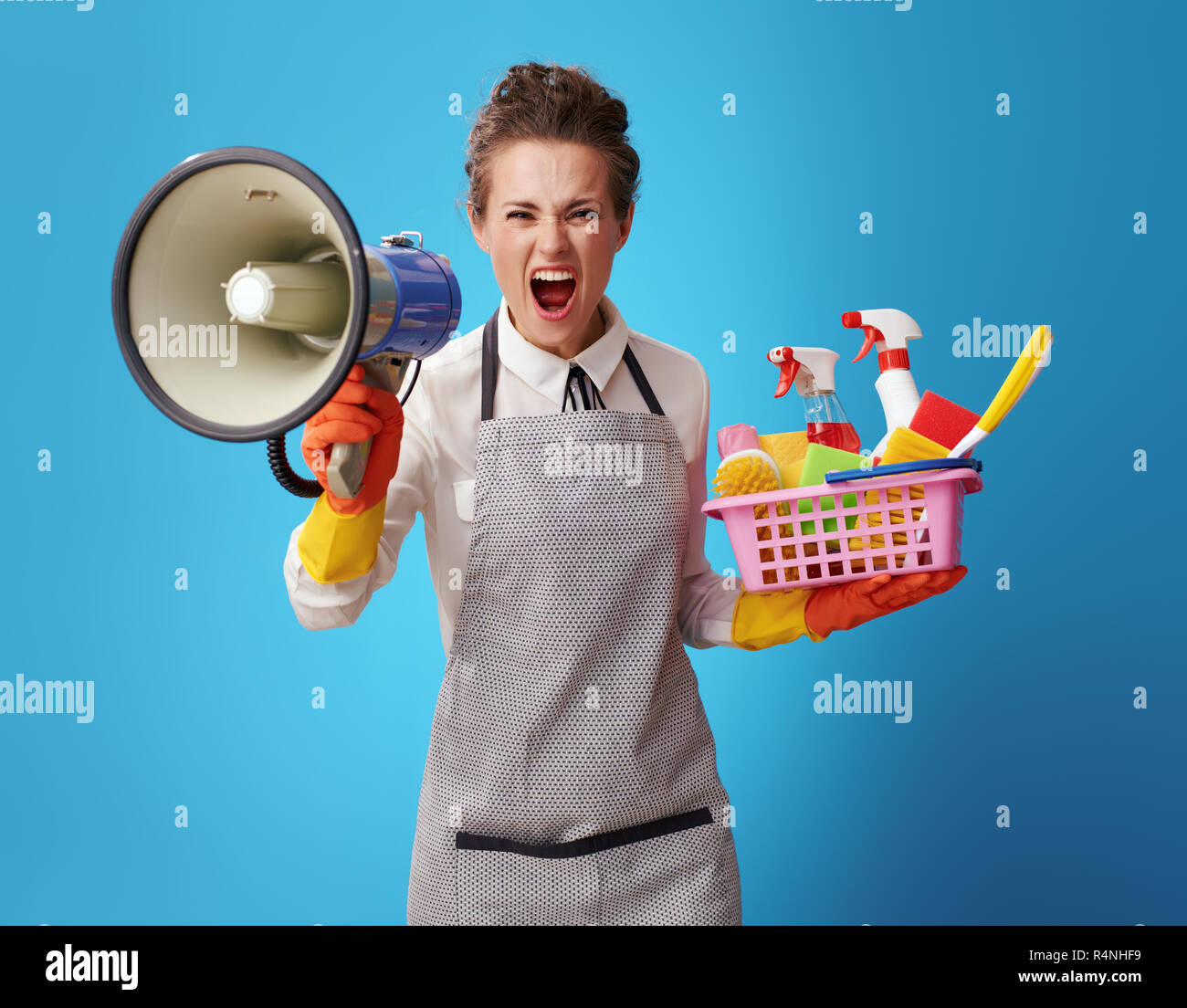 angry young cleaning lady in apron with a basket with cleansers and brushes shouting through a megaphone on blue background. Professional cleaner says Stock Photo