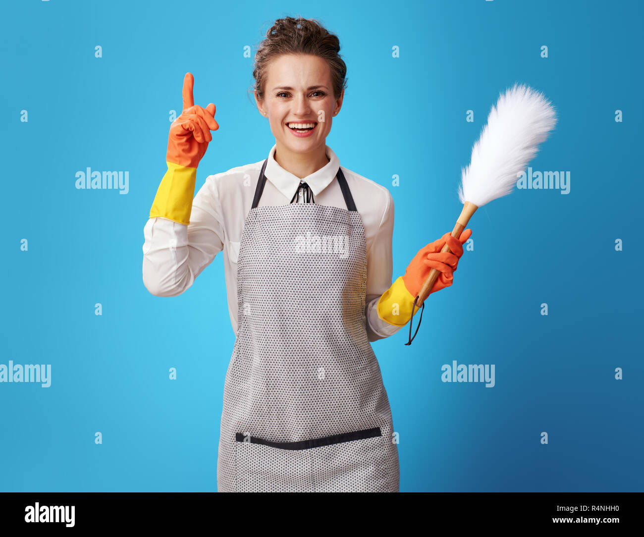 smiling young housemaid in apron with dust cleaning brush got idea against blue background. Where cleaning start? Quality - with the call of professio Stock Photo
