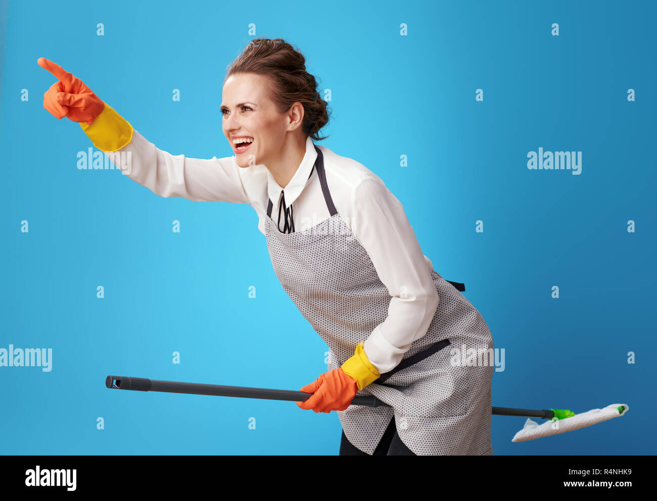 happy scrubwoman in apron pointing at something using mop as a broomstick against blue background. Is it worth spending time on tedious things? there  Stock Photo