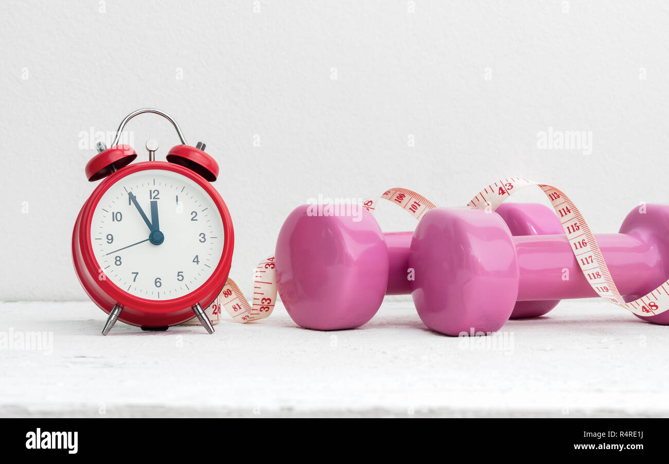 Time to work out, healthy lifestyle and diet concept. Pink dumbbells, alarm clock, and measuring tape on white wood with copy space. Stock Photo