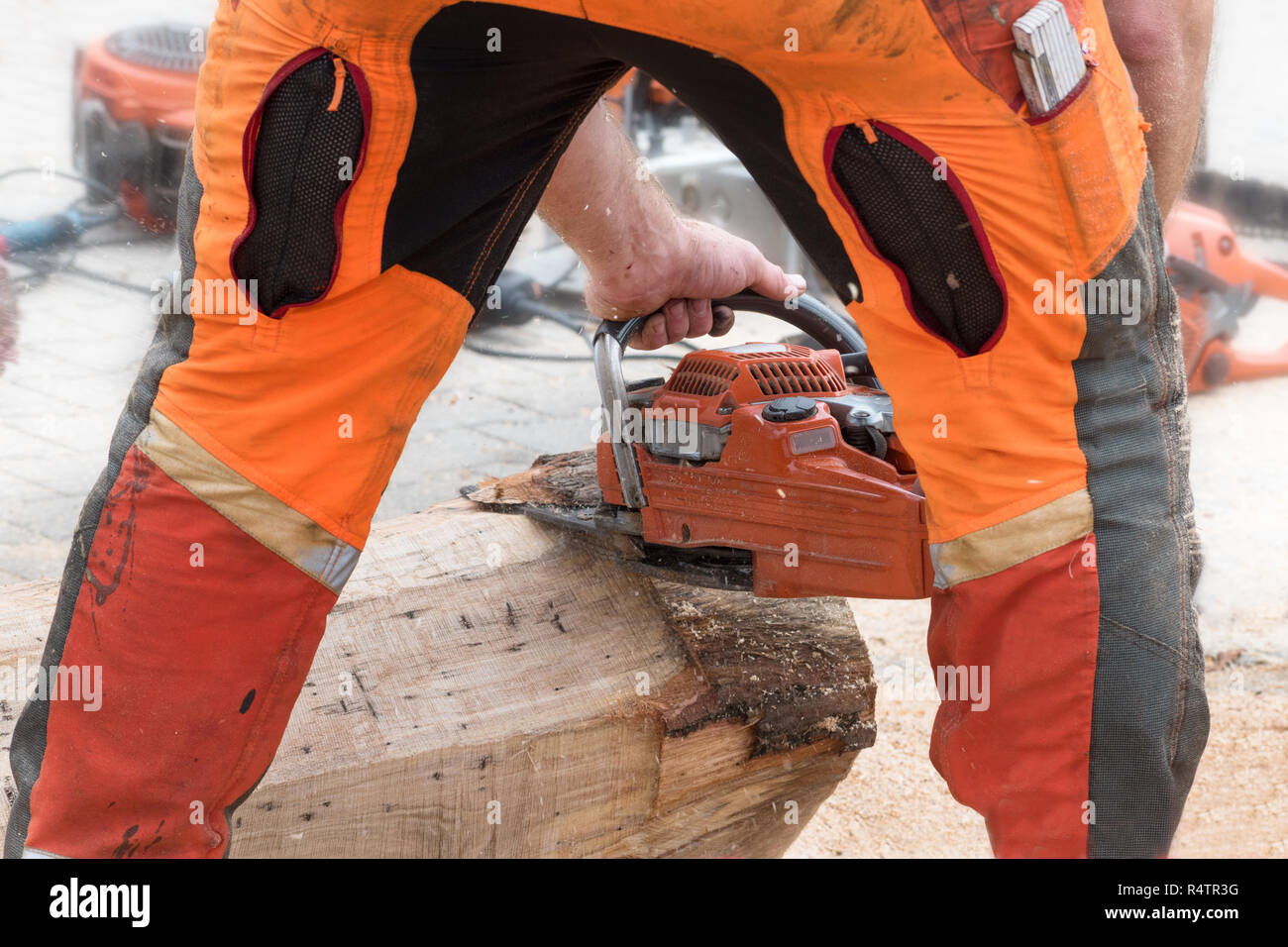 man in orange safety clothing from behind removing the bark from a tree trunk with a chainsaw, view through his legs, selected focus Stock Photo