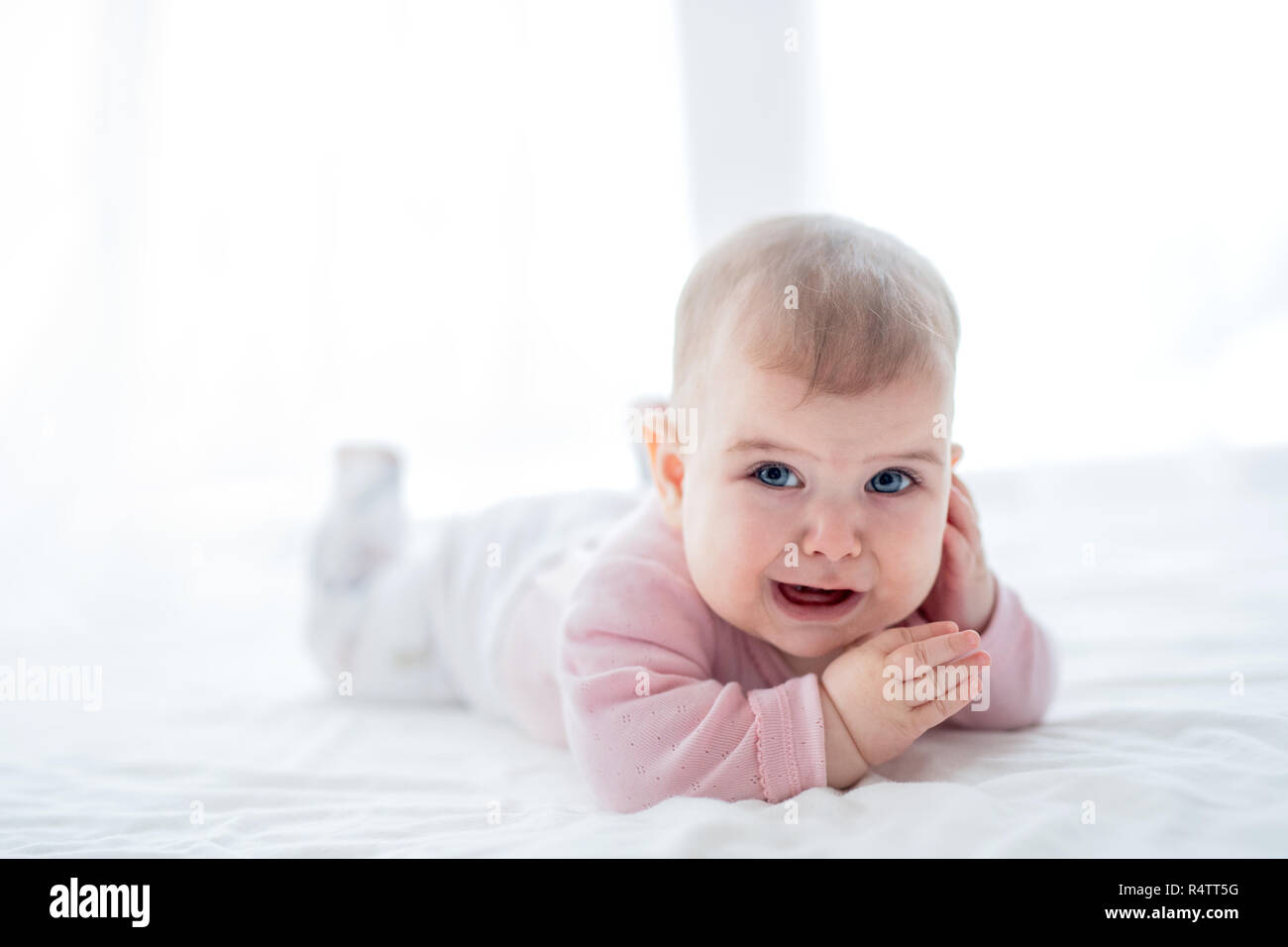 A portrait of unhappy baby girl lying on her tummy on bed indoors. Stock Photo