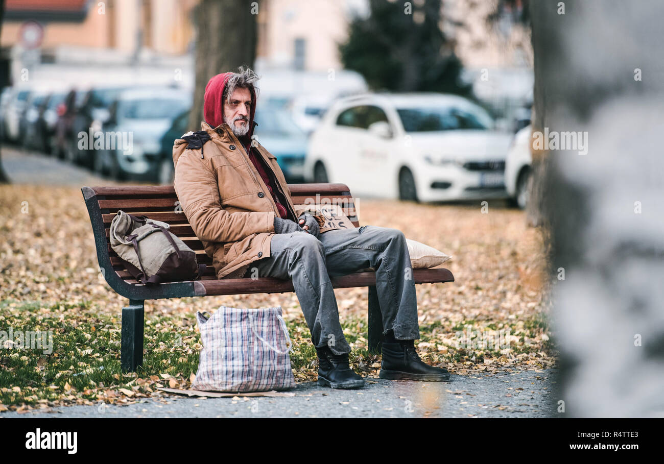 A Homeless Beggar Man With A Bag Sitting On Bench Outdoors In City
