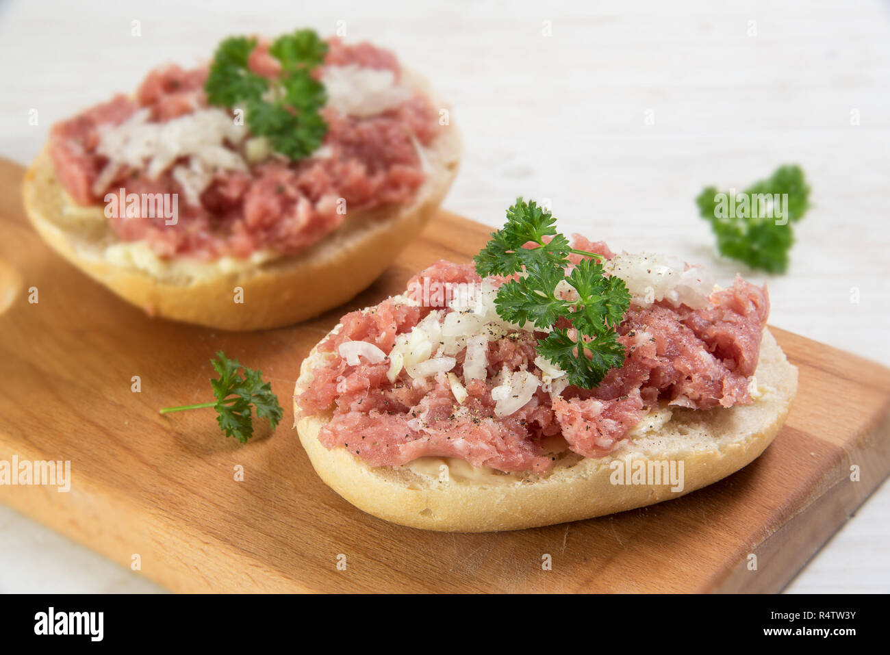 minced pork sausage, typical german mettwurst with onions and parsley garnish on a bun, kitchen board  on a white wooden table, close up with selected Stock Photo
