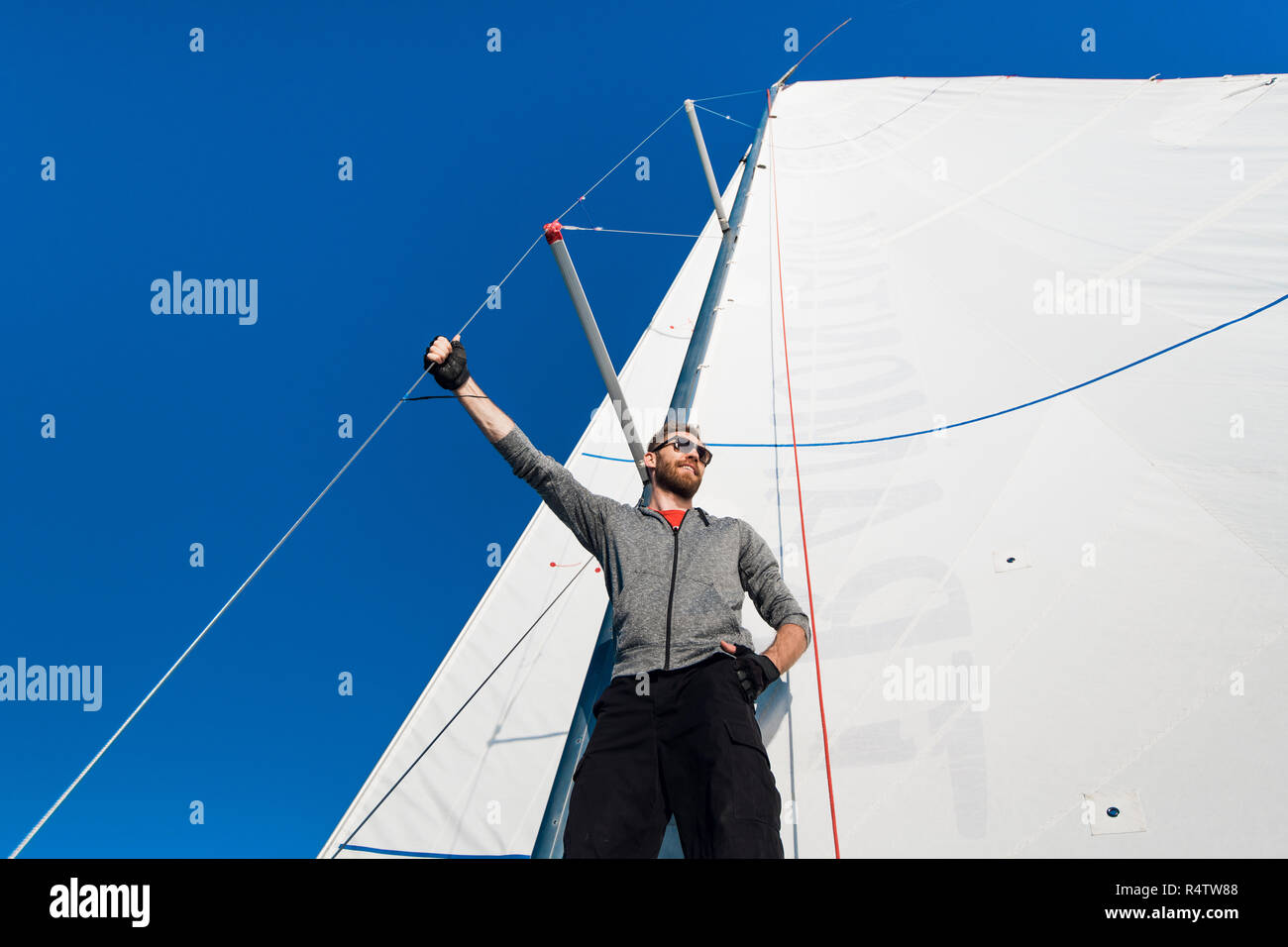Young man stands on yacht boom and looks forward. He holds on mast rope with hand. Young man poses. He wears grey hoody and black pants. Stock Photo