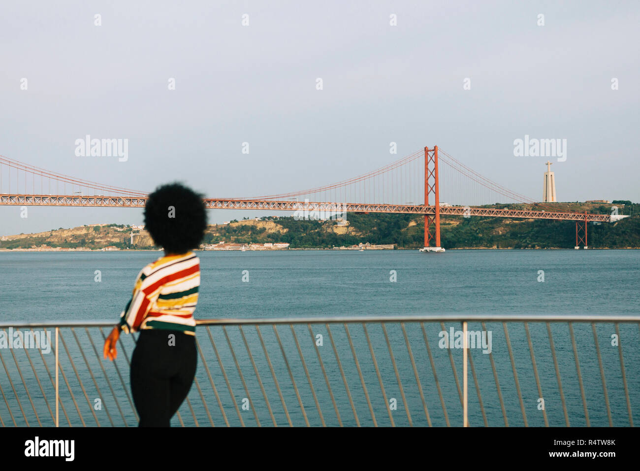 Young woman standing at waterfront, looking at 25 de Abril Bridge and Cristo Rei Statue, Lisbon Stock Photo