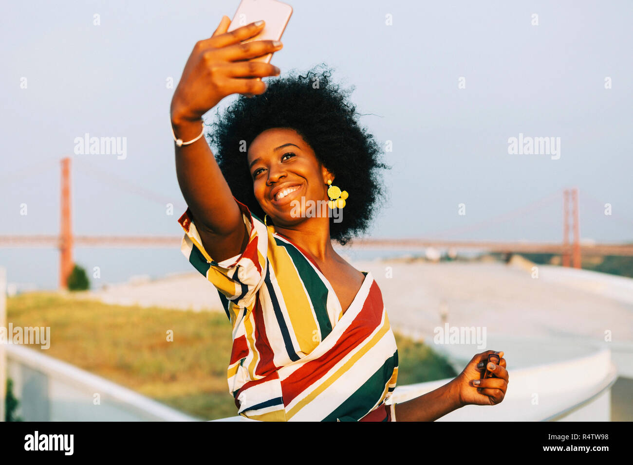 Confident young woman taking selfie with smart phone with 25 de Abril Bridge in background Stock Photo