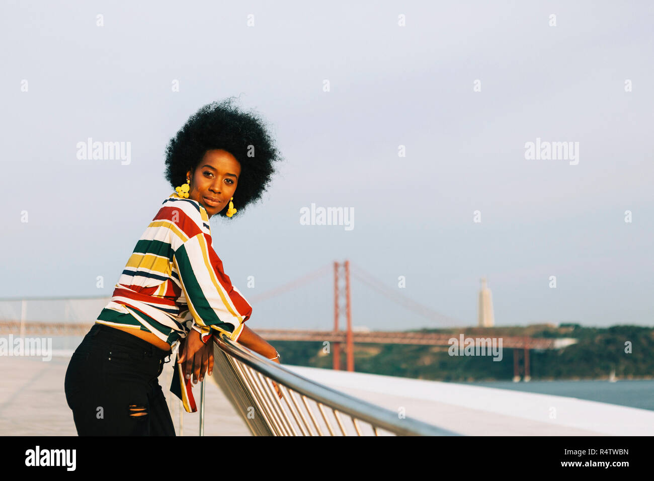 Portrait confident young woman at waterfront with 25 de Abril Bridge in background, Lisbon, Portugal Stock Photo