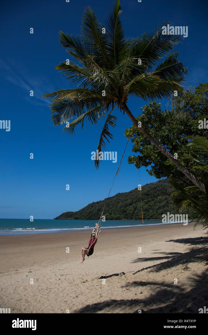 Carefree woman swinging on rope swing below palm tree on tropical beach, Port Douglas, Queensland Stock Photo