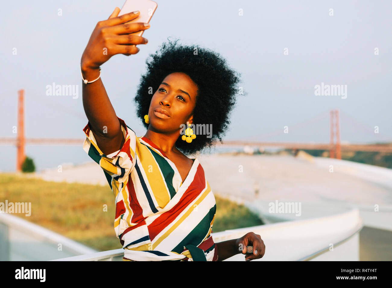 Confident young woman taking selfie with 25 de Abril Bridge in background, Lisbon, Portugal Stock Photo