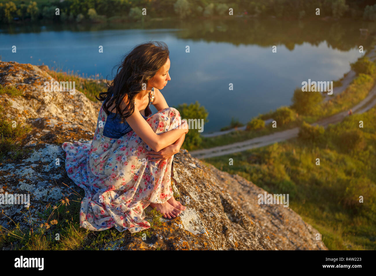 young woman sitting on a cliff overlooking the lake, sad mood, in the evening at sunset. Stock Photo