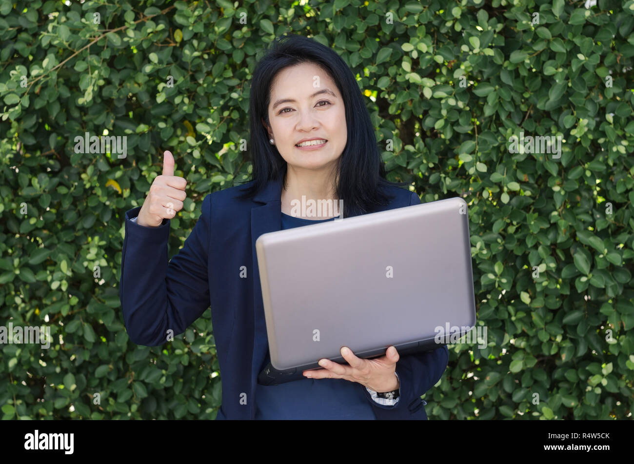 Smiling asian woman holding laptop and showing thumbs up front of green ...