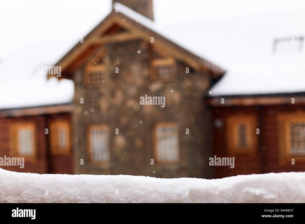 Snow-covered surface for product placement. Blurred house made of wood and stone on background. Snowfall on winter day. Christmas background with wooden cottage or chalet at ski resort. Copyspace. Stock Photo