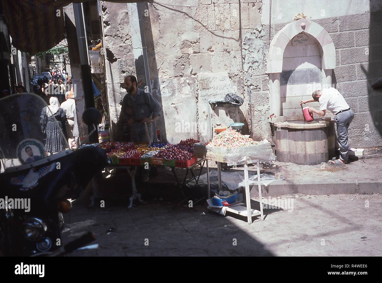 View of a merchant selling fruits and candies in the souk of Damascus, Syria, June 1994. At right, a man draws water from a public stone fountain. At left foreground is a motorcycle with a memorial sticker for Bassel al-Assad, eldest son of President Hafez al-Assad, who died in January 1994. () Stock Photo