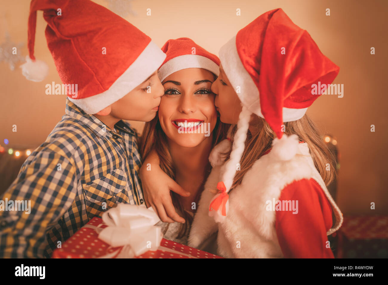 Cute smiling girl and boy give a present to their happy mother for Christmas or New Year. They are kissing and embraced mom. Stock Photo