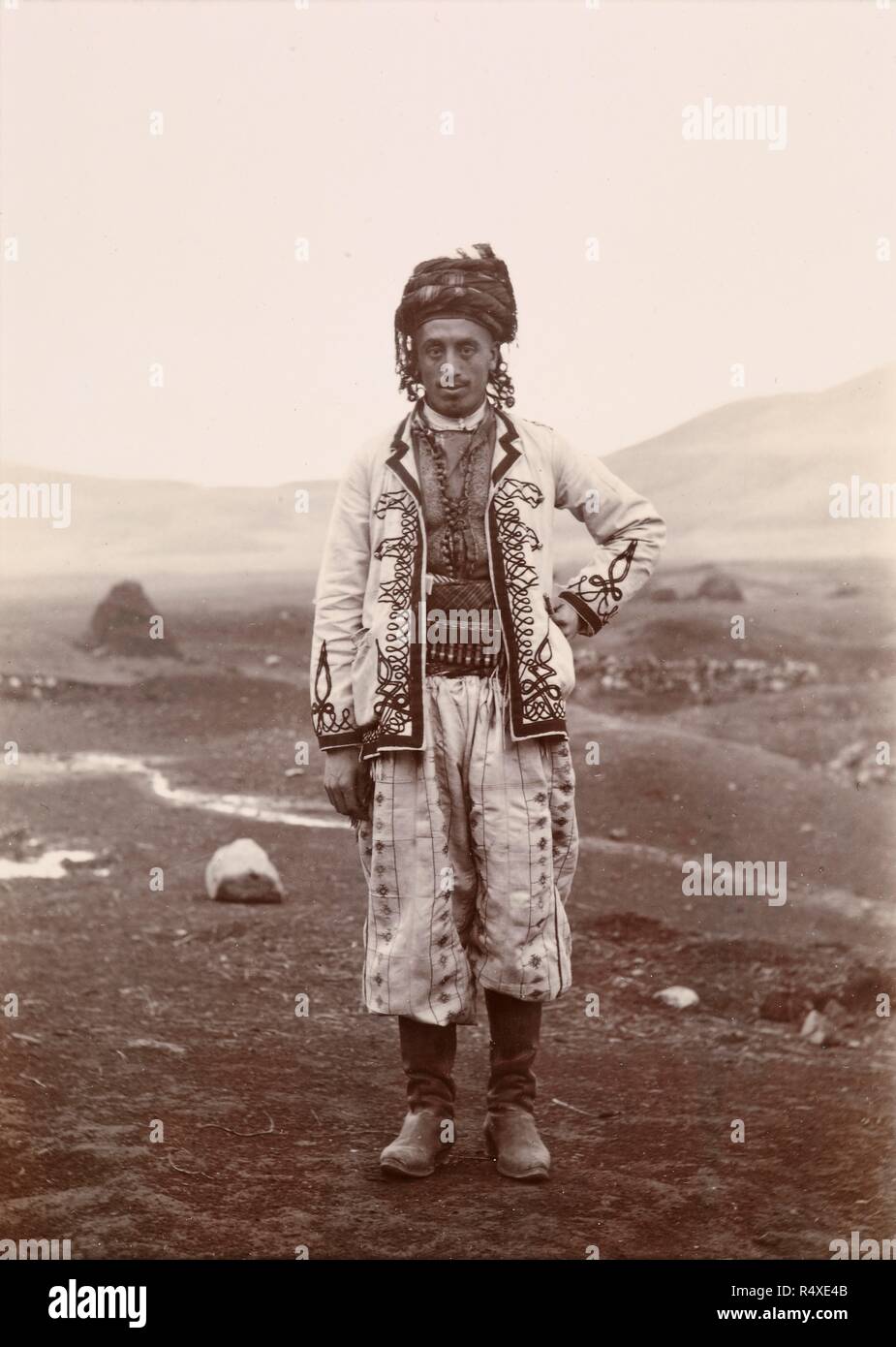 A Kurd in gala dress at village of KÃ¶shk (Sipkanli Kurds). The outskirts of his subterranean village in the background, Autumn 1893. Photographer: Henry Finnis Blosse Lynch, . Source: Photo 430/7 14. Stock Photo