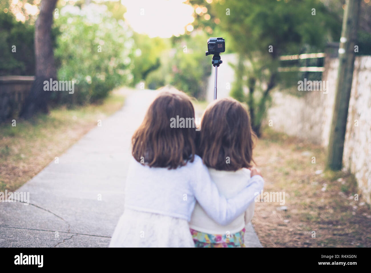 Identical twins sisters walking and taking selfie with action camera. Friends sisters are walking the road and recording themselves with selfie stick  Stock Photo