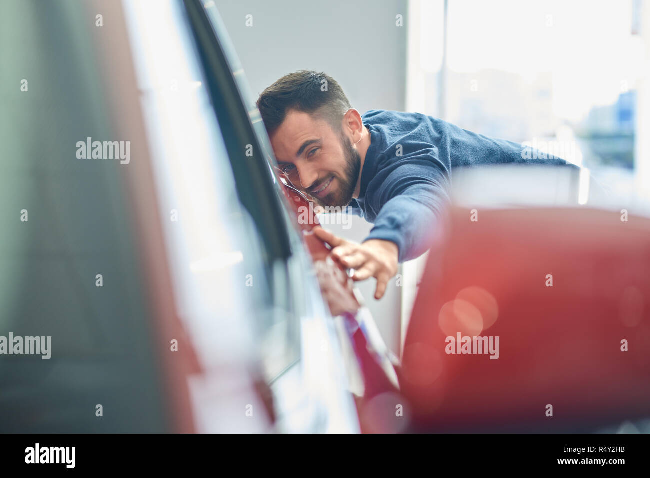 Brunette man with beard coming to salon and buying auto. Young happy client in blue sweater touching new red auto. Attractive driver looking in side mirror and holding his hands on vehicle. Stock Photo