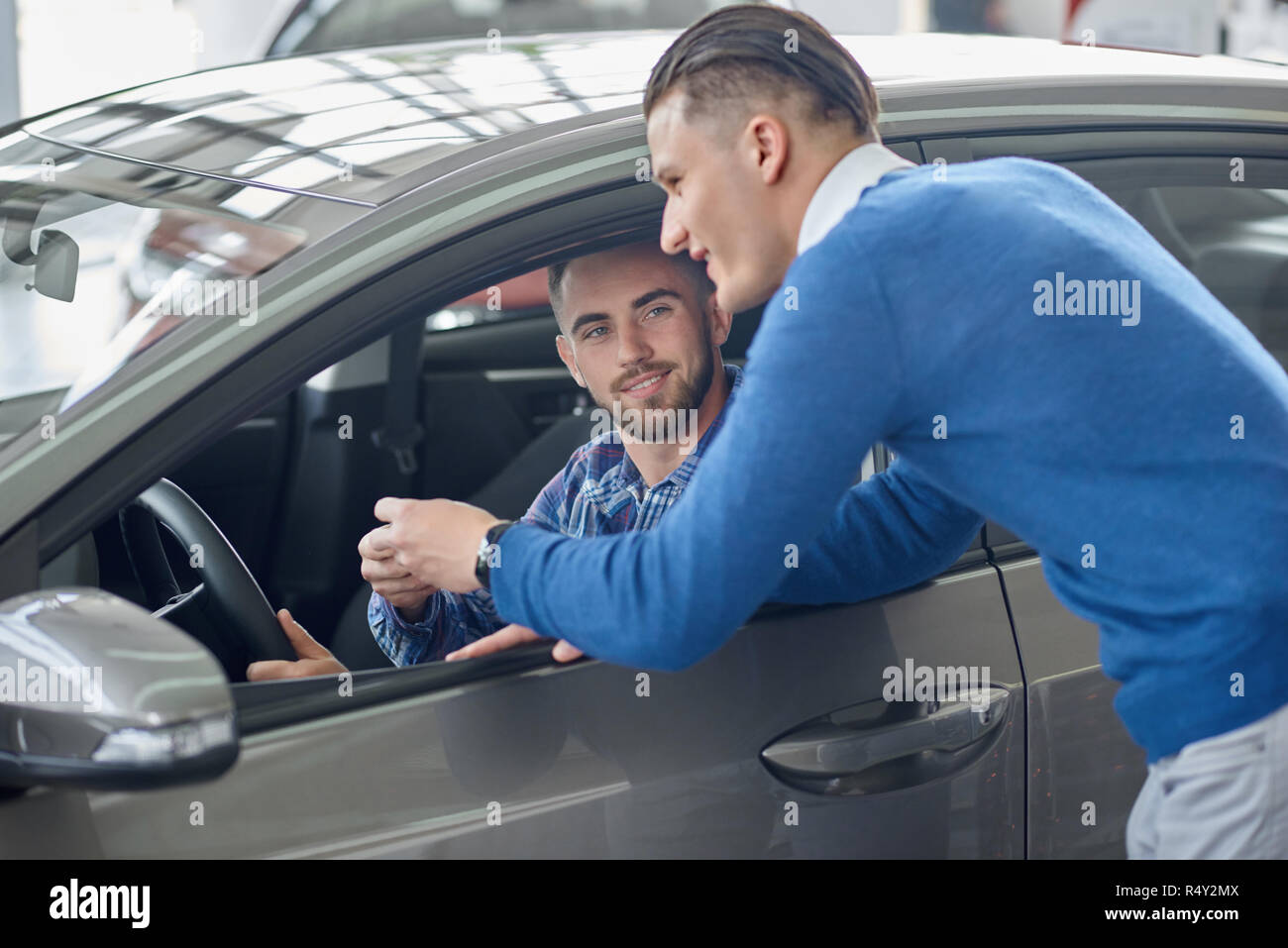 Brunette client with beard sitting in grey car and holding his hand on steering wheel. Menager in blue sweater leaning on front door and talking with customer. Man helping his friend choosing vehicle. Stock Photo