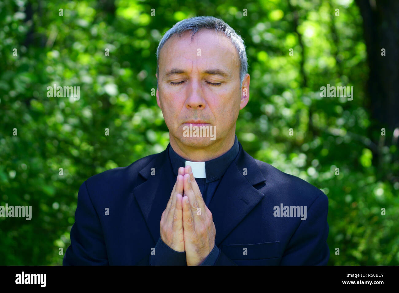 A good looking catholic priest is praying in the forest.  He joins hands. Eyes closed. Stock Photo