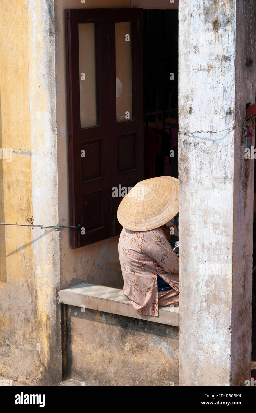 Elderly woman in traditional Vietnamese clothing sitting on stone wall  wearing straw coolie hat in Hoi An Old Town, Quang Nam Province, Vietnam Stock Photo