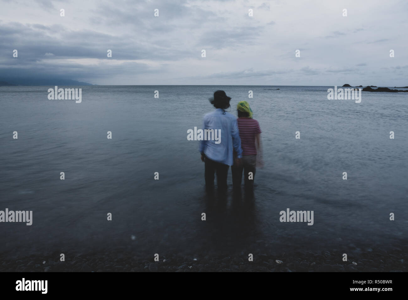 Fully clothed couple, blurred, stand in shallow water, with their back to viewer, as they look out to sea, long exposure, Taiwan. Soft filtered images Stock Photo