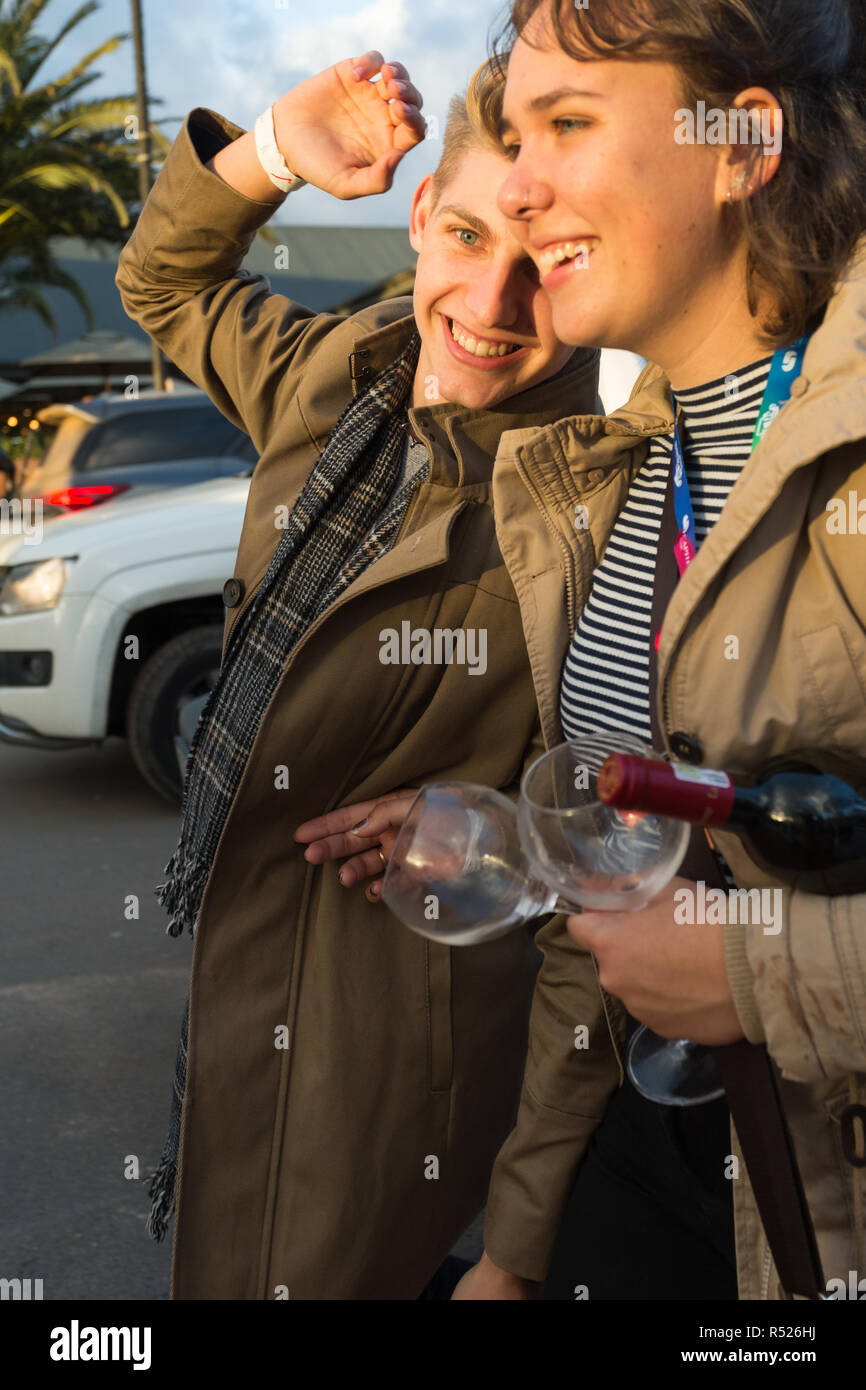 Young boy and girl or couple between 18 to 22 years of age walking in the street carrying a bottle of red wine and two empty glasses smiling and happy Stock Photo