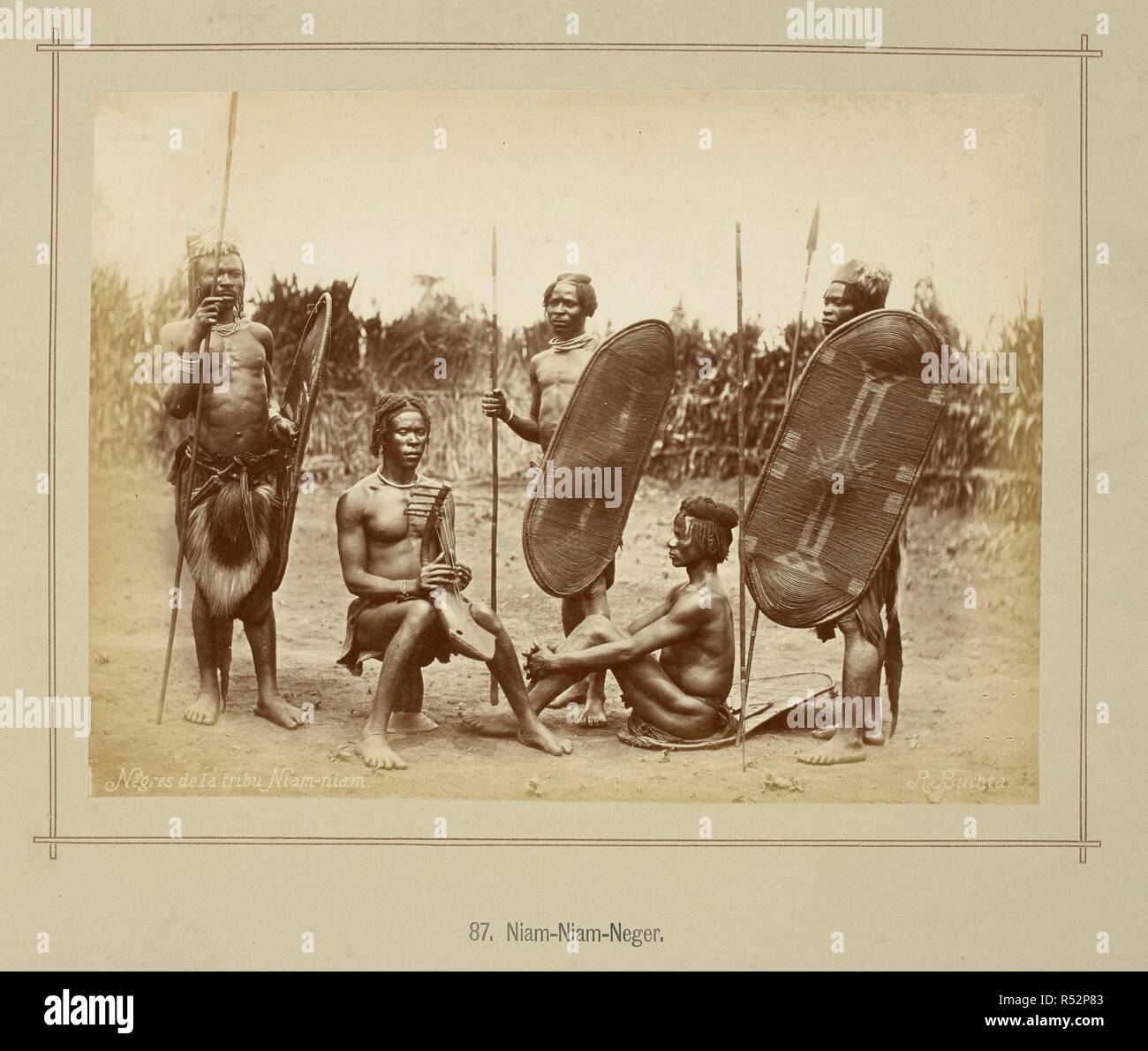 A group of five Zande men, three standing and two seated, one on a carved stool playing a harp, the other on a shield. The standing men are all carrying a spear and shield, two wearing grass-woven hats and one wearing a monkey skin in front of his barkcloth.  Southern Sudan. Die oberen Nil-LaÌˆnder. Volkstypen und Landschaften. Dargestellt in 160 Photographien. Nach der Natur aufgenommen von R. Buchta. Mit einer Einleitung von Dr. Robert Hartmann. Berlin, 1881. Source: 1789.a.13 plate 87. Language: German. Author: Buchta, Richard. Stock Photo