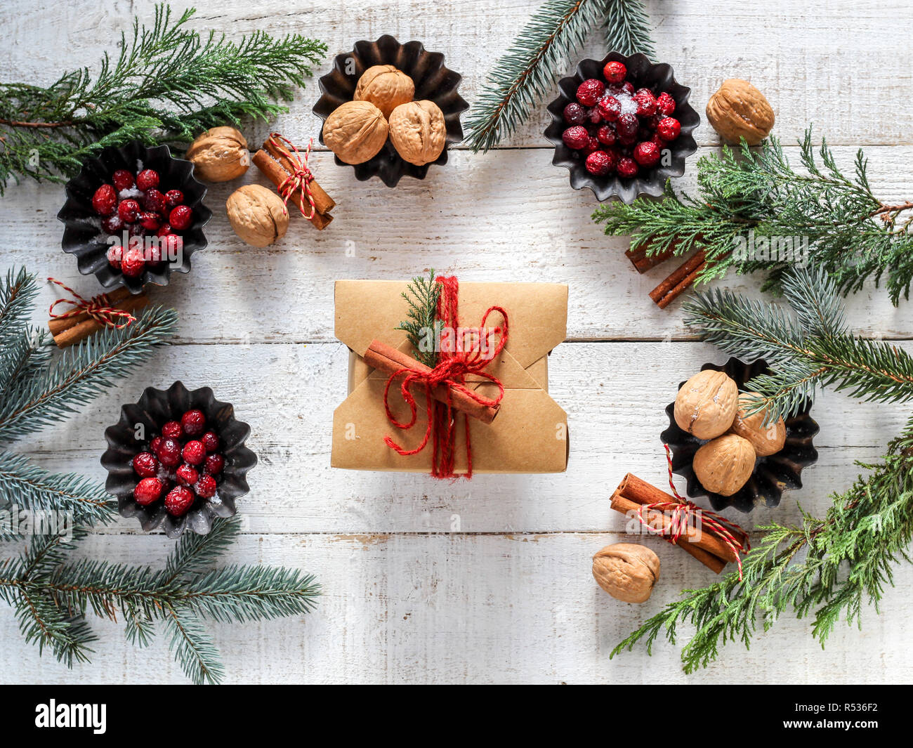 Festive mood flat lay with a gift box in the middle surrounded by evergreens, red berries, nuts and cinnamon sticks on a white wooden background Stock Photo