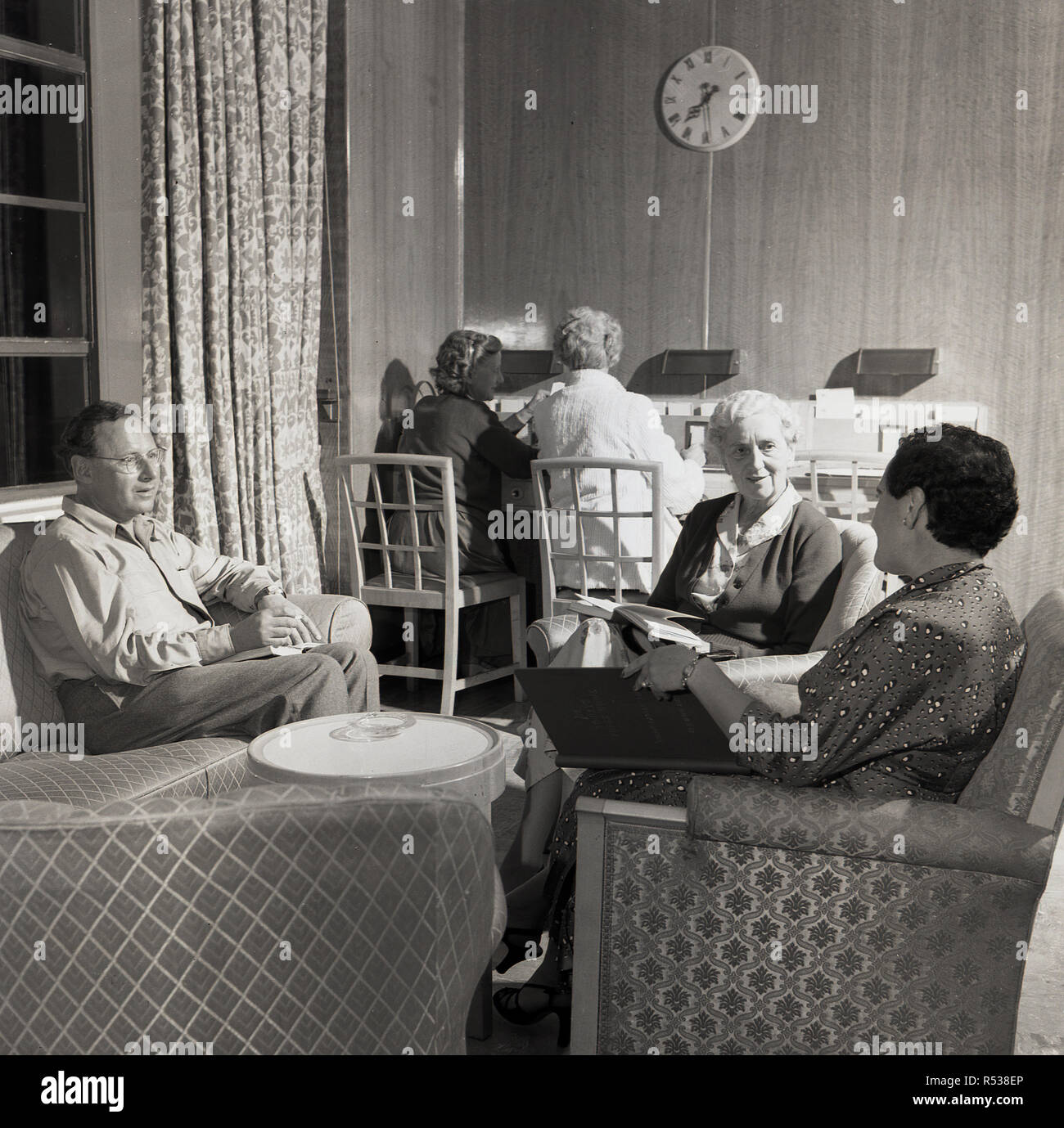 1950s, historical, early evening and several adult passengers onboard a union-castle steamship sitting together relaxing in comfortable chairs inside a ship's lounge. Stock Photo