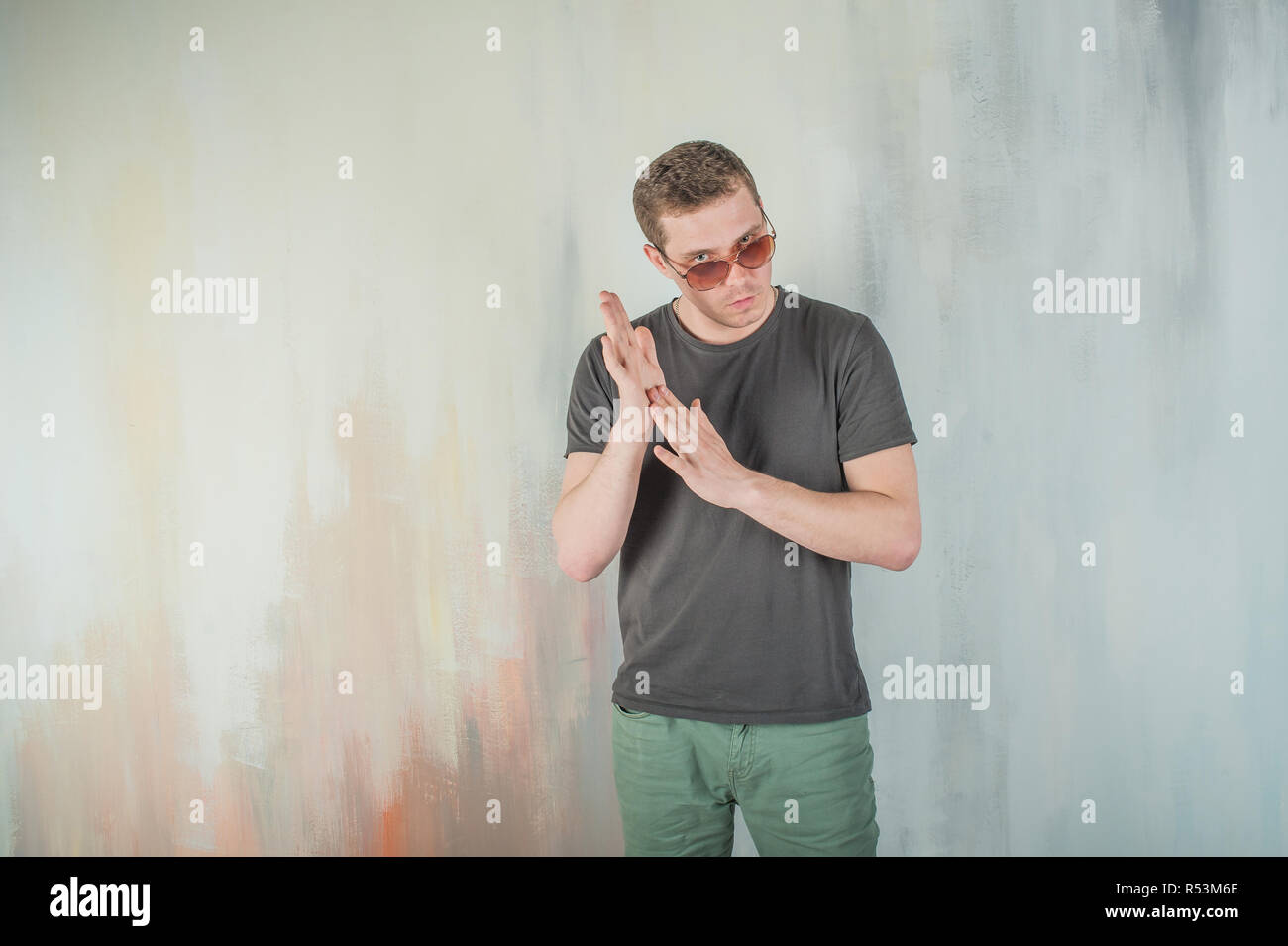 studio photography young brutal guy. man in sunglasses, T-shirt, jeans on a background colorful painted walls. his hands clasped palm to palm rubbing them Stock Photo