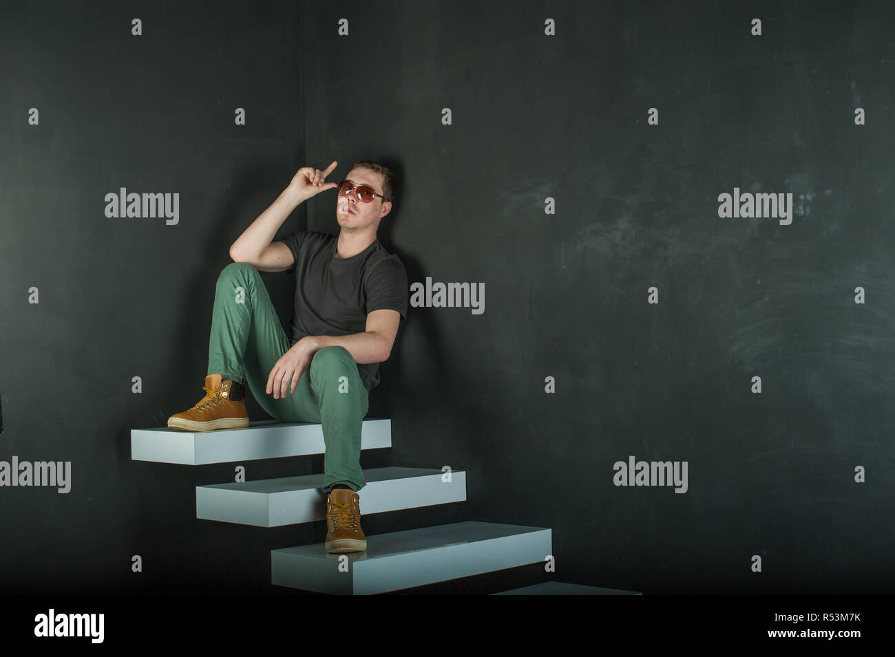 studio photography young brutal guy. man in sunglasses, T-shirt, jeans and high boots on a background of black wall on the white steps. sitting on the steps Stock Photo