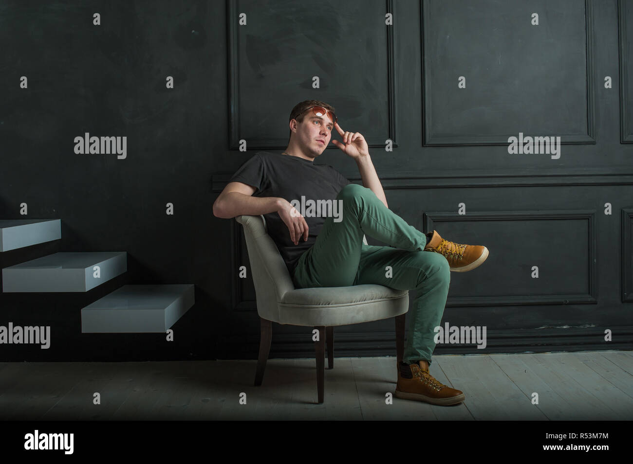 studio photography young brutal guy. man in sunglasses, T-shirt, jeans and high boots on a background of black wall, He is sitting in a white armchair throne. Stock Photo