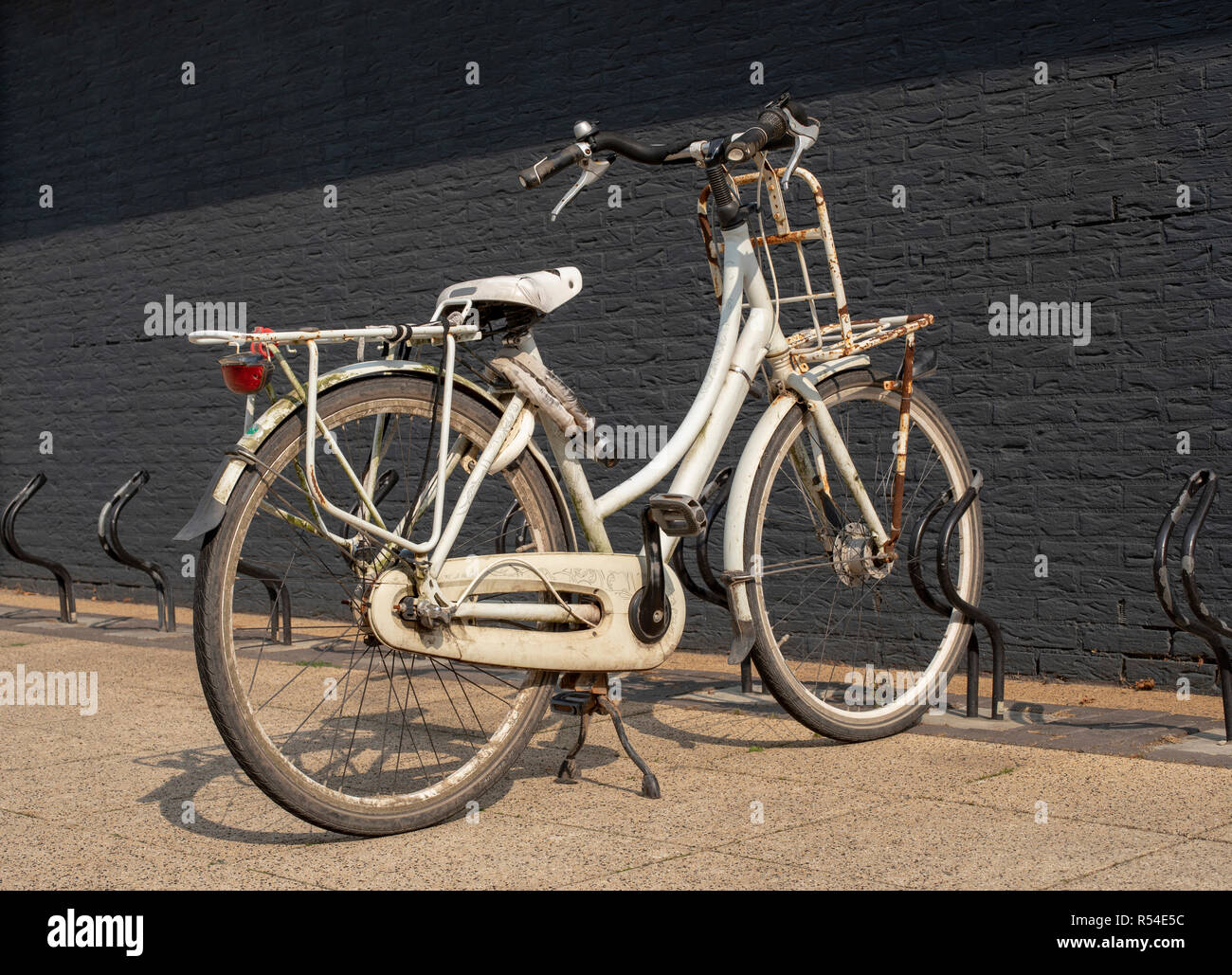 Old rusty grunge vintage white bike in bicycle rack in front of a black painted brick wall. Stock Photo
