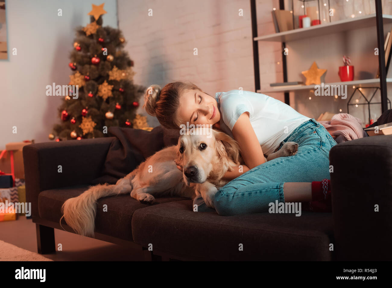 beautiful content young blonde woman on couch hugging golden retriever dog at christmas time Stock Photo