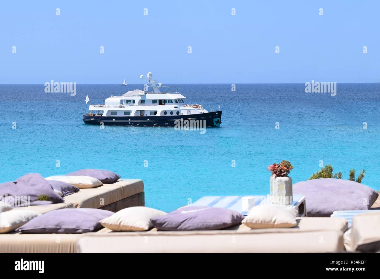 luxurious blue yacht anchored in turquoise waters off the coast of formentera,with benches and cushions on the shore in the foreground Stock Photo
