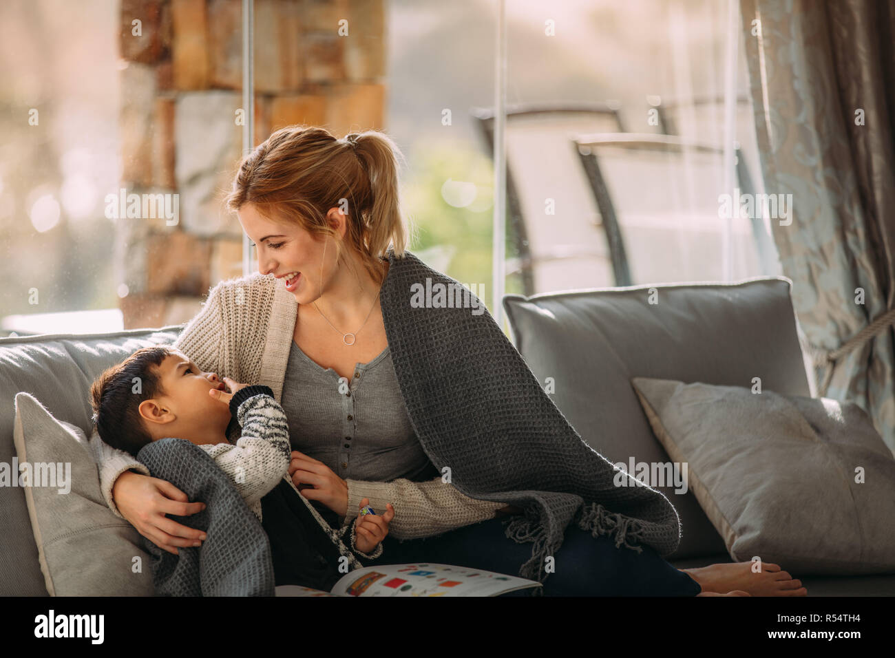 Little boy touching his mouth and looking at his mother sitting on sofa. Mother and son having fun time together at home. Stock Photo