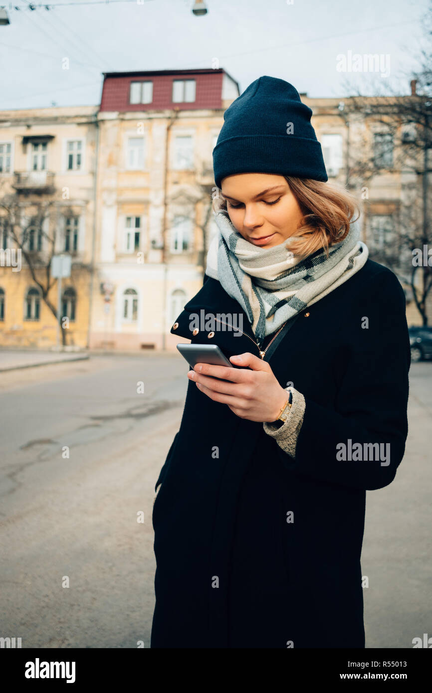 Female smiling looking at mobile device on winter street. Young caucasian woman wearing wool coat, scarf and hat using smart phone in city. Stock Photo