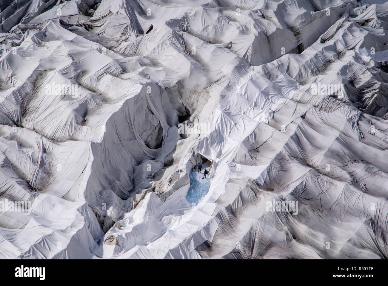 Two persons can be seen at the entrance of a glacier ice cave at the mouth of the Rhone Glacier in Switzerland. The ice cave and the surrounding glacier are covered every year with huge sheets of fleece blankets to slow down the inevitable melting of the glacier due to warmer climate. A popular tourist attraction since the 1870s, the glacier is now disappearing fast. With higher mean temperatures and lack of precipitation, the Rhone Glacier is shrinking dramatically. An exceptionally snowy winter was not enough to offset the extreme summer, which caused glaciers to lose 2.5% of their volume, t Stock Photo